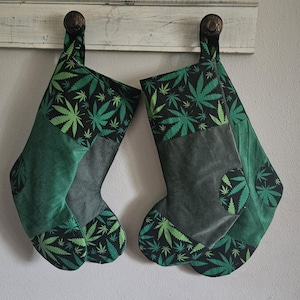 May include: Two Christmas stockings with a cannabis leaf pattern. The stockings are green, black, and gray, with a corduroy texture. The cannabis leaf pattern is in shades of green on a black background.