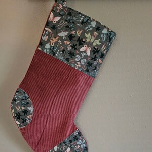May include: A red and gray Christmas stocking with a woodland-themed pattern featuring butterflies, mushrooms, and leaves. The stocking has a red corduroy cuff and a gray fabric top with a loop for hanging.