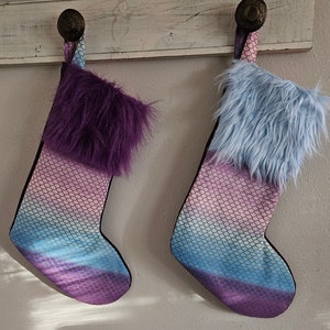 May include: Two Christmas stockings with a mermaid scale pattern in shades of purple, pink, and blue. Each stocking has a fluffy faux fur cuff in either purple or light blue. The stockings are hanging on a white wooden surface.