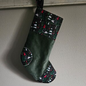 May include: A green Christmas stocking with a white and black pattern of stylized Christmas trees. The stocking has a corduroy cuff.