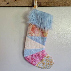 May include: A patchwork Christmas stocking with a blue faux fur cuff. The stocking is made of various fabrics with floral and geometric patterns in shades of pink, yellow, blue, and white.