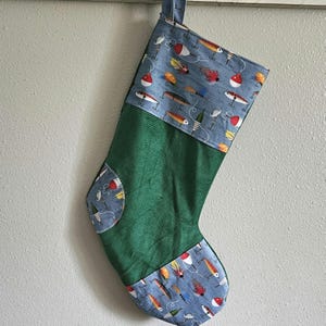 Peut inclure: Une chaussette de Noël verte et bleue avec un motif de leurres de pêche. La chaussette est en velours côtelé et possède une boucle en haut pour l'accrocher.