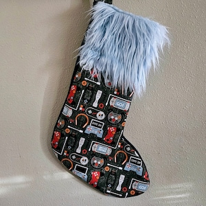 May include: A Christmas stocking with a black background, patterned with red, blue, and white video game controllers, consoles, and accessories. The top of the stocking is trimmed with light blue faux fur.