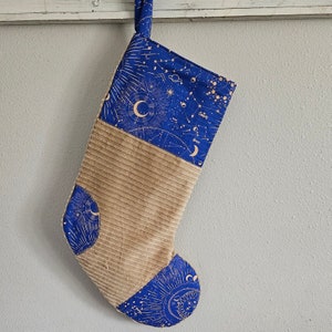 May include: A blue and brown Christmas stocking with a celestial pattern featuring stars, moons, and planets. The stocking is made of corduroy and has a gold metallic print.