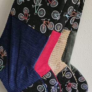 May include: A Christmas stocking with a patchwork design. The stocking features a black fabric with colorful bicycle illustrations, navy blue corduroy, red, tan, and gray fabrics. The stocking is designed for the holiday season.