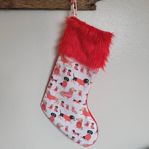 May include: A red and white Christmas stocking with a fluffy red cuff. The stocking is decorated with a pattern of dachshunds wearing sweaters.
