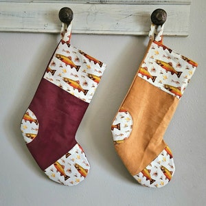 Peut inclure: Deux chaussettes de Noël sur le thème de la pêche. Une chaussette est bordeaux avec un revers blanc et l'autre est beige avec un revers blanc. Les deux chaussettes présentent un motif de truites et de mouches de pêche.