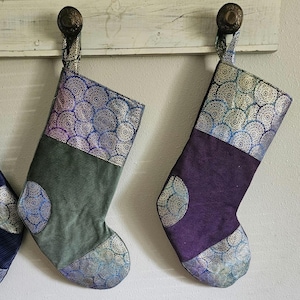 May include: Two Christmas stockings with a decorative design. One is green, the other is purple. Both have a patterned top and toe, with a matching circular design on the body. They hang from a white wooden shelf.