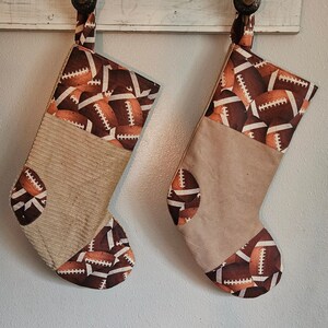 May include: Two Christmas stockings with a football theme. The top and bottom of the stockings are patterned with brown and white footballs. The middle section is a tan corduroy fabric. Each stocking has a loop for hanging.