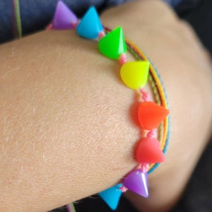 May include: A rainbow colored bracelet with a series of pointed, teardrop-shaped beads. The bracelet is made with colorful string and is worn on a person's wrist.