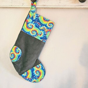 May include: A Christmas stocking with a gray corduroy body and tie-dye fabric accents in blue, green, yellow, and red. The stocking is hanging from a white wooden hook.