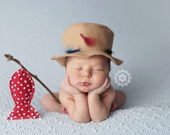 May include: A sleeping baby wearing a brown felt hat with feathers and a red and white polka dot fish toy.