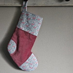 May include: A handmade Christmas stocking with a burgundy corduroy body and a flamingo and leaf patterned top and accents. The stocking is hanging from a white wooden hook against a neutral wall.