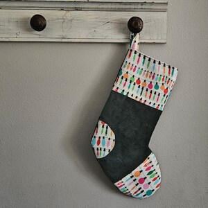May include: A black and white stocking with a pattern of colorful light bulbs. The stocking is hanging on a white wooden shelf with two black hooks.