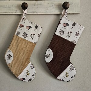 May include: Two brown corduroy Christmas stockings with white fabric panels featuring a mushroom design. The stockings are hung on a white wooden surface.