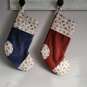 Peut inclure: Deux chaussettes de Noël en tissu de velours côtelé avec un panneau en tissu crème orné d'un motif de mouches de pêche. Une chaussette est bleue et l'autre est rouge.