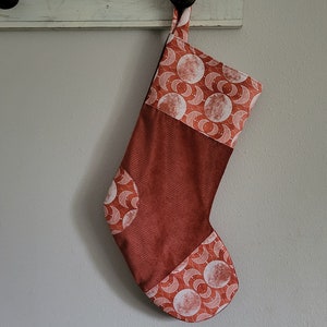 May include: A Christmas stocking with a red and white celestial pattern. The stocking is made of a textured, reddish-brown fabric with a white and red moon and crescent design. It hangs from a white wooden hook.