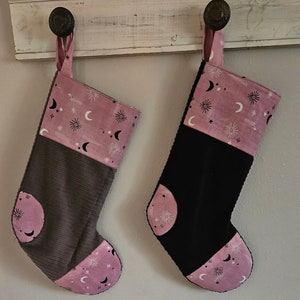 May include: Two Christmas stockings hanging from a white wooden shelf. One stocking is gray corduroy, the other is black. Both have a pink top and toe with a celestial pattern of moons and stars.