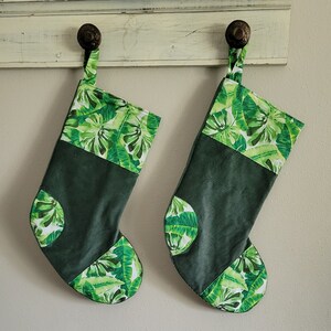 May include: Two green Christmas stockings with a tropical leaf pattern. The stockings are made of a dark green corduroy fabric with a white and green leaf pattern at the top and bottom. They are hanging from a white shelf.