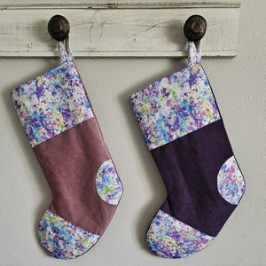 May include: Two Christmas stockings with a colorful splatter paint design. One stocking is mauve with a matching accent circle, the other is purple. Both have a white top with a colorful splatter design and are hanging from a white wooden shelf.