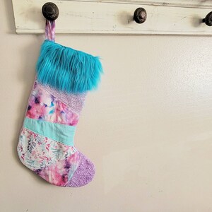 May include: A colorful patchwork Christmas stocking with a blue faux fur cuff. The stocking is hanging on a white hook.