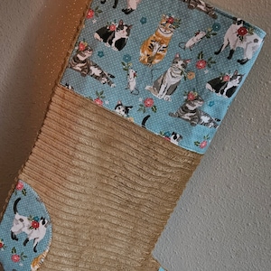 Può includere: Un calzino di Natale in velluto a coste marrone con una parte superiore in tessuto blu e bianco con un motivo di gatti e fiori. Il calzino ha un anello in cima per appenderlo.