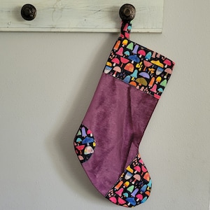 May include: A purple velvet stocking with a colorful mushroom pattern on the cuff. The stocking is hung on a white wooden surface.