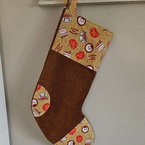 May include: A brown Christmas stocking with a corduroy texture. The top and bottom sections feature a repeating pattern of ramen bowls, noodles, and text. The stocking is hanging from a black hook.