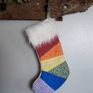 May include: A handmade Christmas stocking with a rainbow design. The stocking features sections of red chevron, orange polka dots, yellow, green floral, blue, and purple dot fabric. The top is trimmed with white faux fur.