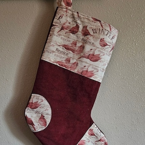 May include: A red and white Christmas stocking with a winter theme. The stocking features a white fabric with a pattern of red cardinals, branches, and the words "Winter Garden", "Rejoice", and "Happy Heart Dream". The stocking is trimmed with a red corduroy fabric.