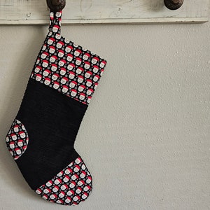May include: A Christmas stocking with a black ribbed body and a patterned top and toe featuring Santa Claus faces on a black background. The stocking is hanging from a white wooden rack.