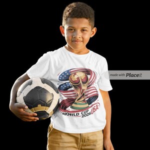 May include: A young person wearing a white t-shirt with a graphic of the World Cup trophy, American flag, and the text "WORLD CUP 2026 USA". They are holding a black and white soccer ball.