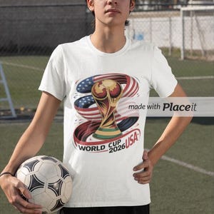 May include: White t-shirt featuring a graphic of the World Cup trophy with the American flag and the text "World Cup 2026 USA". The person is holding a soccer ball.