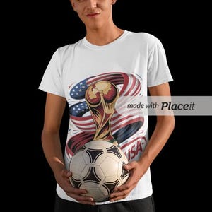 May include: White t-shirt with a soccer ball and a graphic of the FIFA World Cup trophy, the American flag, and the word "USA". The shirt is worn by a person.