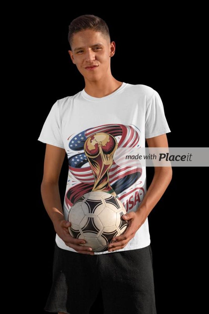 May include: White t-shirt with a soccer ball and a graphic of the FIFA World Cup trophy, the American flag, and the word "USA". The shirt is worn by a person.