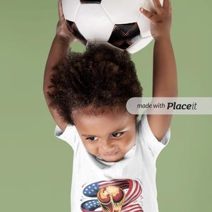 May include: A child holding a soccer ball above their head. The child is wearing a white t-shirt with a graphic of the World Cup trophy, the American flag, and the text "WORLD CUP 2026 USA". The soccer ball is white with black pentagons.