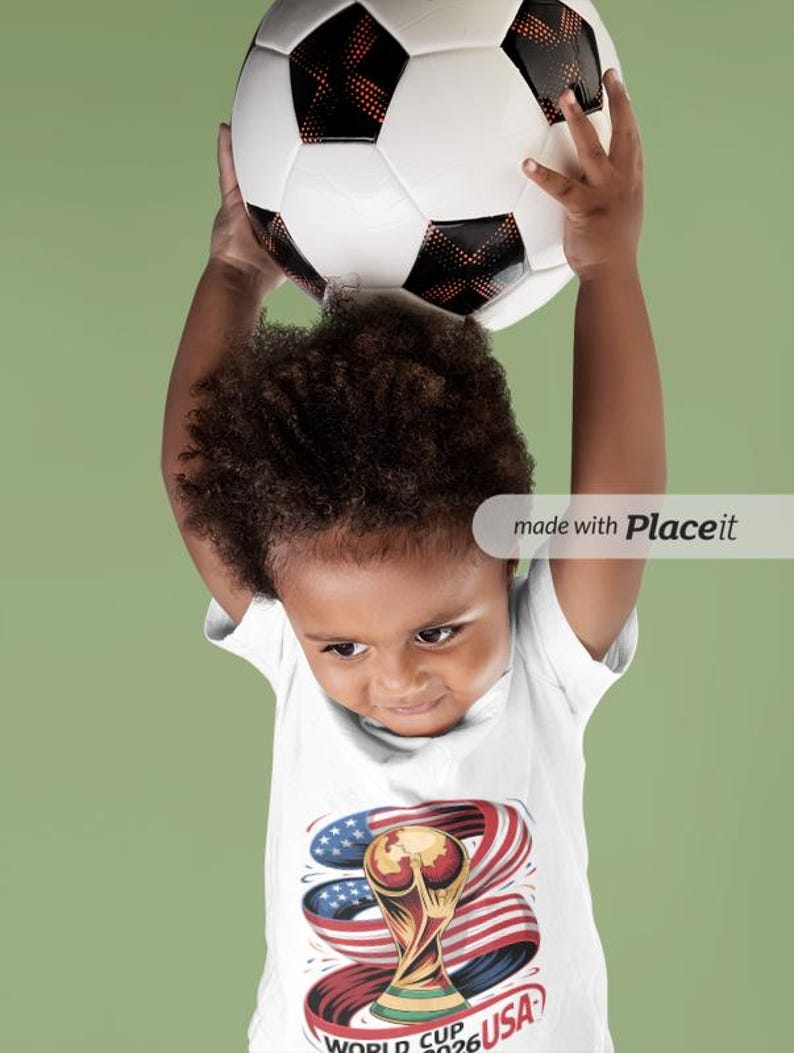 May include: A child holding a soccer ball above their head. The child is wearing a white t-shirt with a graphic of the World Cup trophy, the American flag, and the text "WORLD CUP 2026 USA". The soccer ball is white with black pentagons.