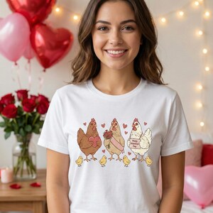 May include: White t-shirt featuring a whimsical illustration of three chickens, each with a unique Valentine's Day theme. One chicken holds a heart, another carries flowers, and the third displays a sign that reads "Happy Valentine's Day". Small yellow chicks are at the bottom.