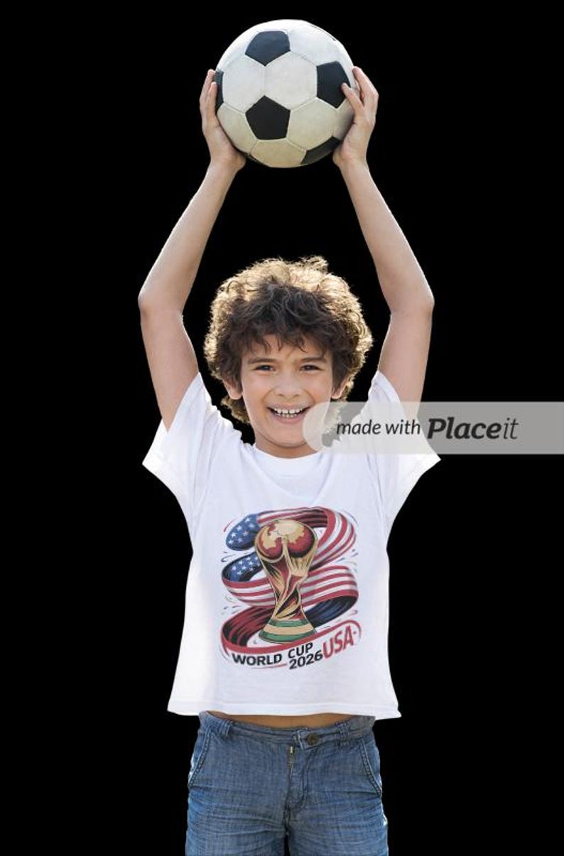 May include: A young person holds a soccer ball overhead, smiling. They wear a white t-shirt with a graphic of the World Cup trophy, the American flag, and the text "World Cup 2026 USA". The background is black.