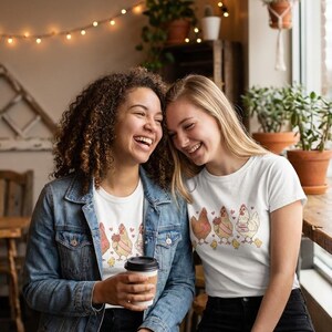 May include: Two women wearing white t-shirts with a chicken graphic. The shirts feature a row of cartoon chickens with hearts and flowers. One woman wears a denim jacket and holds a coffee cup. The other woman is smiling.