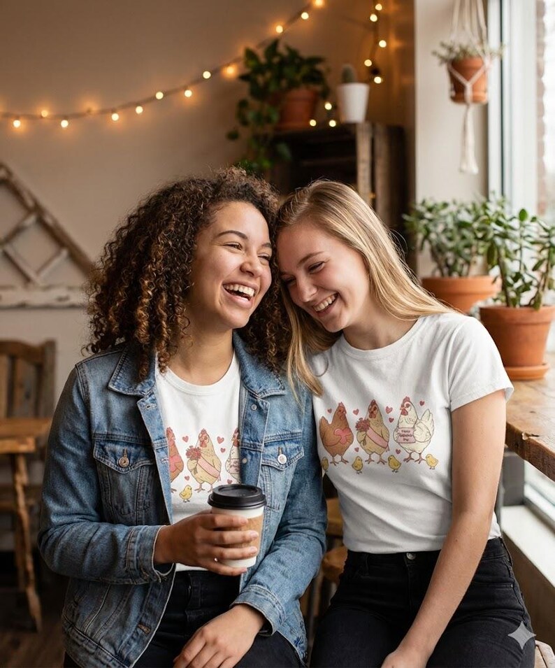 May include: Two women wearing white t-shirts with a chicken graphic. The shirts feature a row of cartoon chickens with hearts and flowers. One woman wears a denim jacket and holds a coffee cup. The other woman is smiling.