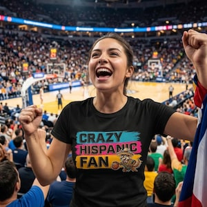 Puede incluir: Una camiseta negra con el texto "CRAZY HISPANIC FAN" en letras de colores. La camiseta presenta un mapache de dibujos animados con un sombrero y un balón de baloncesto. La persona que lleva la camiseta anima en un partido de baloncesto.