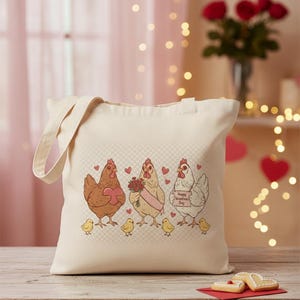May include: Cream-colored tote bag featuring a whimsical illustration of three chickens, each holding a heart, flowers, and a sign that says "Happy Valentine's Day." The chickens are accompanied by chicks. The bag has a simple, rectangular shape with handles.
