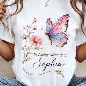 May include: A white t-shirt featuring a watercolor-style graphic of a pink and blue butterfly and a floral stem. Below the butterfly, the text reads In Loving Memory of Sophia in a combination of serif and elegant script fonts on the front.