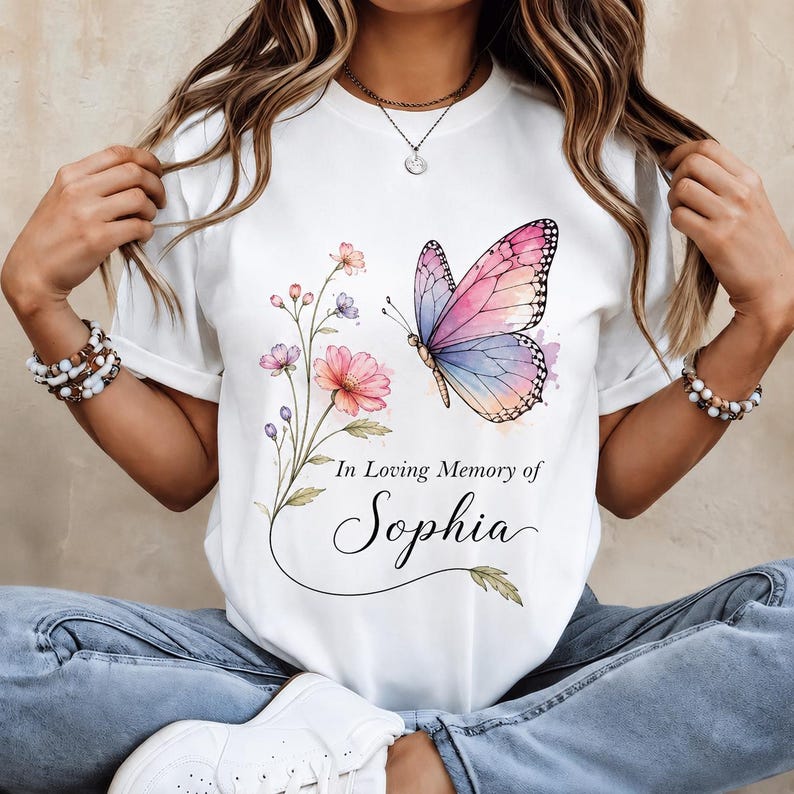 May include: A white t-shirt featuring a watercolor-style graphic of a pink and blue butterfly and a floral stem. Below the butterfly, the text reads In Loving Memory of Sophia in a combination of serif and elegant script fonts on the front.