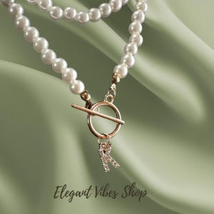 Può includere: Una collana di perle a due fili con chiusura color oro e un ciondolo con iniziale. Il ciondolo è una lettera "R" tempestata di strass. La collana è mostrata su uno sfondo di tessuto verde. Il testo "Elegant Vibes Shop" è in basso.