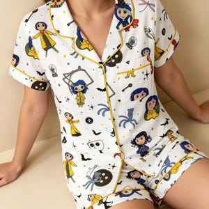 May include: A white short-sleeve pajama set with a button-down shirt and matching shorts, featuring a repeating pattern of cartoon characters, black buttons, bats, and stars. The set is accented with yellow piping along the collar, pocket, and cuffs.