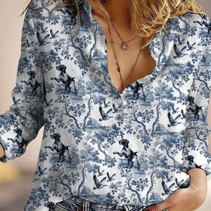 May include: A white button-down shirt with a blue toile pattern featuring dogs and birds. The long-sleeved shirt has a classic collar and a relaxed fit, suitable for casual wear. The shirt is paired with blue jeans.