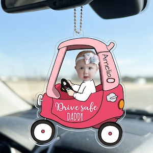 Könnte beinhalten: Ein rosa, auto-förmiger Autoanhänger mit einem Babyfoto. Der Anhänger trägt den Namen "Amelia" und den Text "Drive safe DADDY". Er hängt am Rückspiegel eines Autos.