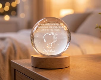May include: A clear glass sphere resting on a circular light-colored wooden base. The sphere features an etched design of a stethoscope shaped like a heart with floral accents, the name Sophia, and the text: Never underestimate the difference you make and the lives you touch.